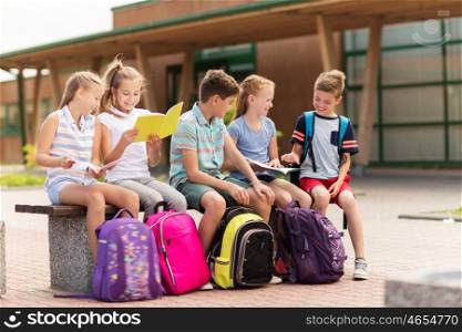 primary education, friendship, childhood, communication and people concept - group of happy elementary school students with backpacks and notebooks sitting on bench outdoors