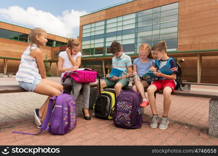 primary education, friendship, childhood, communication and people concept - group of happy elementary school students with backpacks and notebooks sitting on bench and doing homework outdoors