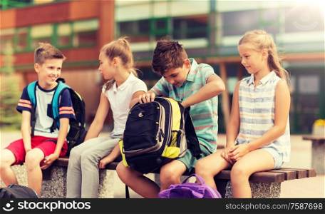primary education, friendship, childhood, communication and people concept - group of happy elementary school students with backpacks sitting on bench outdoors. group of elementary school students with backpacks