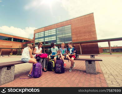 primary education, friendship, childhood, communication and people concept - group of happy elementary school students with backpacks and notebooks sitting on bench and doing homework outdoors. group of happy elementary school students outdoors