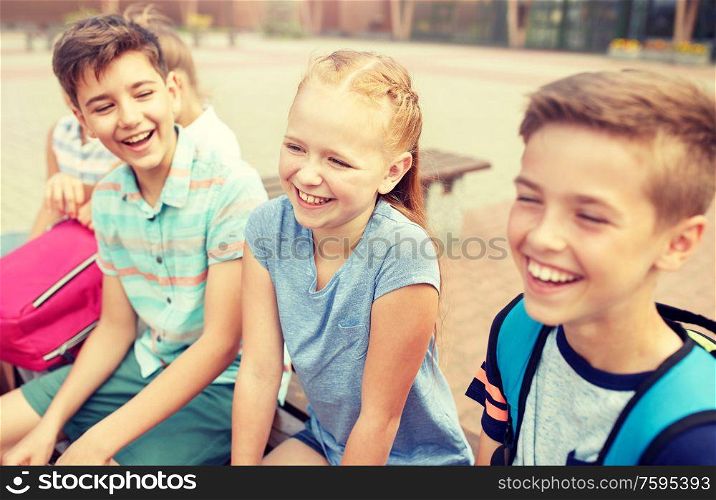 primary education, friendship, childhood, communication and people concept - group of happy elementary school students with backpacks sitting on bench and talking outdoors. group of happy elementary school students talking