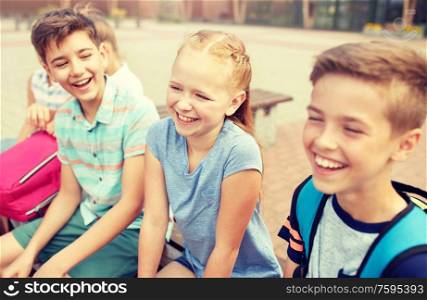 primary education, friendship, childhood, communication and people concept - group of happy elementary school students with backpacks sitting on bench and talking outdoors. group of happy elementary school students talking