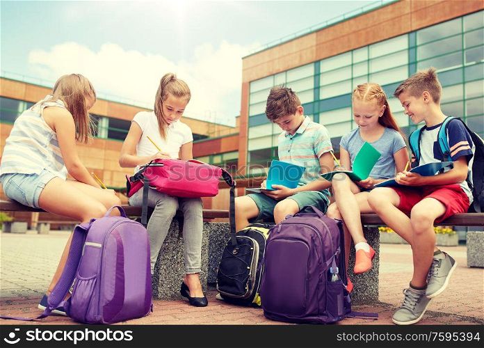 primary education, friendship, childhood, communication and people concept - group of happy elementary school students with backpacks and notebooks sitting on bench and doing homework outdoors. group of happy elementary school students outdoors
