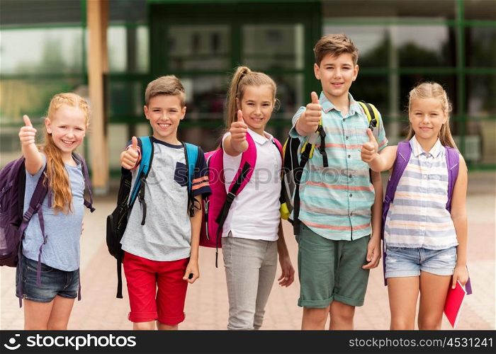 primary education, friendship, childhood, gesture and people concept - group of happy elementary school students with backpacks showing thumbs up outdoors