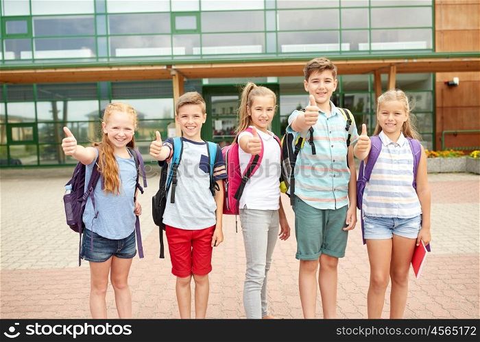 primary education, friendship, childhood, gesture and people concept - group of happy elementary school students with backpacks showing thumbs up outdoors