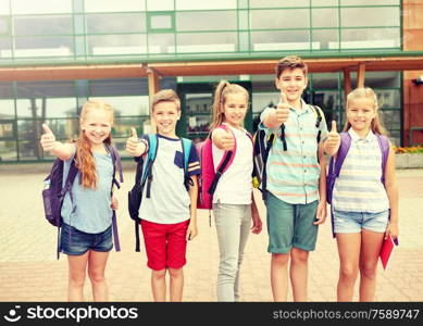 primary education, friendship, childhood, gesture and people concept - group of happy elementary school students with backpacks showing thumbs up outdoors. happy elementary school students showing thumbs up