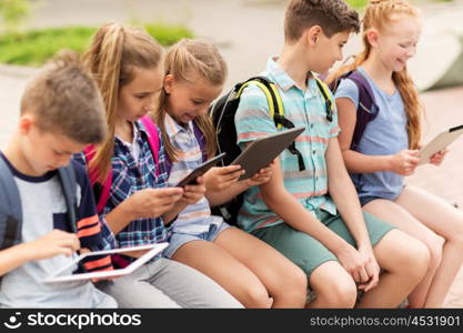 primary education, friendship, childhood, technology and people concept - group of happy elementary school students with backpacks sitting on bench and talking outdoors