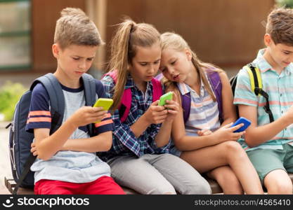 primary education, friendship, childhood, technology and people concept - group of happy elementary school students with smartphones and backpacks sitting on bench outdoors. elementary school students with smartphones