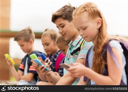 primary education, friendship, childhood, technology and people concept - group of happy elementary school students with smartphones and backpacks sitting on bench outdoors. elementary school students with smartphones