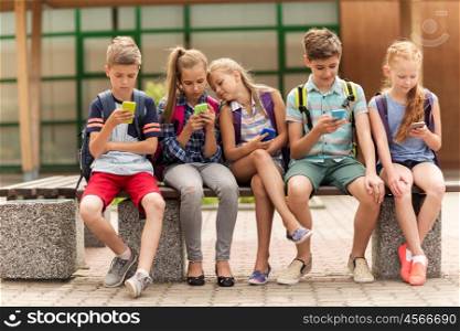 primary education, friendship, childhood, technology and people concept - group of happy elementary school students with smartphones and backpacks sitting on bench outdoors