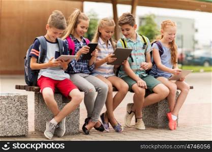 primary education, friendship, childhood, technology and people concept - group of happy elementary school students with backpacks sitting on bench and talking outdoors