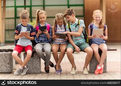 primary education, friendship, childhood, technology and people concept - group of happy elementary school students with backpacks sitting on bench and talking outdoors