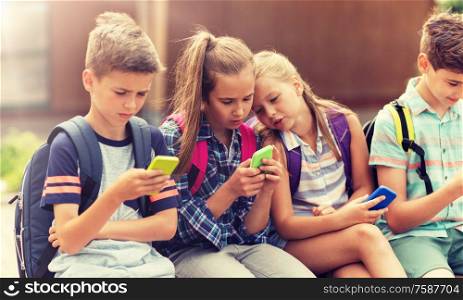 primary education, friendship, childhood, technology and people concept - group of happy elementary school students with smartphones and backpacks sitting on bench outdoors. elementary school students with smartphones
