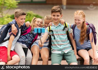 primary education, technology, friendship, childhood and people concept - group of elementary school students with backpacks sitting on bench and taking picture by smartphone on selfie stick outdoors