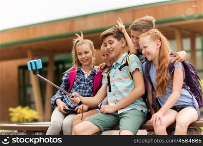 primary education, technology, friendship, childhood and people concept - group of elementary school students with backpacks sitting on bench and taking picture by smartphone on selfie stick outdoors