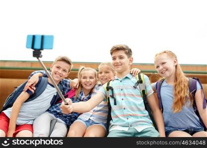 primary education, technology, friendship, childhood and people concept - group of elementary school students with backpacks sitting on bench and taking picture by smartphone selfie stick outdoors