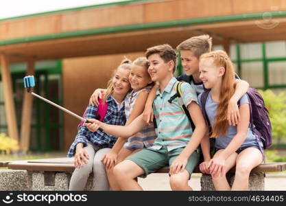 primary education, technology, friendship, childhood and people concept - group of elementary school students with backpacks sitting on bench and taking picture by smartphone on selfie stick outdoors