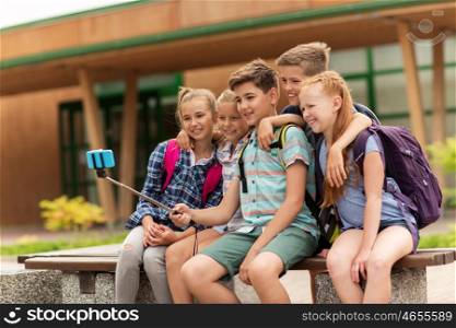 primary education, technology, friendship, childhood and people concept - group of elementary school students with backpacks sitting on bench taking picture by smartphone on selfie stick outdoors