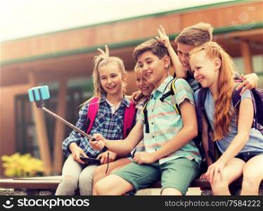 primary education, technology, friendship, childhood and people concept - group of elementary school students with backpacks sitting on bench and taking picture by smartphone on selfie stick outdoors. happy elementary school students taking selfie