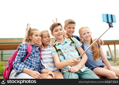primary education, technology, friendship, childhood and people concept - group of happy school students with backpacks sitting on bench and taking picture by smartphone on selfie stick outdoors