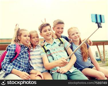primary education, technology, friendship, childhood and people concept - group of happy school students with backpacks sitting on bench and taking picture by smartphone on selfie stick outdoors. happy elementary school students taking selfie