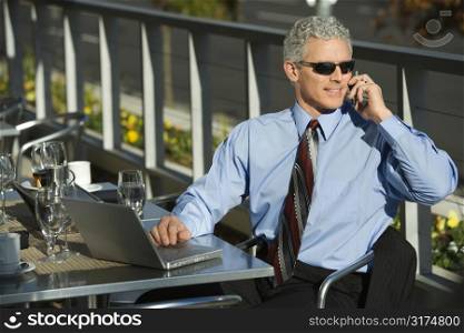 Prime adult Caucasian man in suit sitting at patio table ouside with laptop and talking on cellphone.