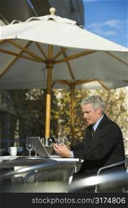 Prime adult Caucasian man in suit sitting at patio table outside with laptop and dialing cellphone.