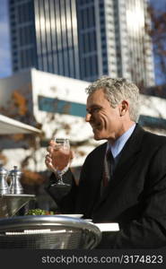 Prime adult Caucasian man in suit sitting outside drinking water at patio table in urban setting.