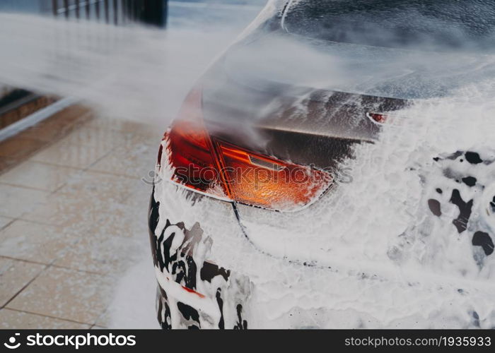 Process of removing dirt from car exterior surface with high pressure washer at self-serve cleaning station outdoors, rinsing off chemicals and white soap suds from vehicle at carwash center. Washing car exterior surface with high pressure washer at self-serve cleaning station outdoors
