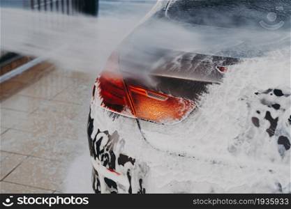 Process of removing dirt from car exterior surface with high pressure washer at self-serve cleaning station outdoors, rinsing off chemicals and white soap suds from vehicle at carwash center. Washing car exterior surface with high pressure washer at self-serve cleaning station outdoors
