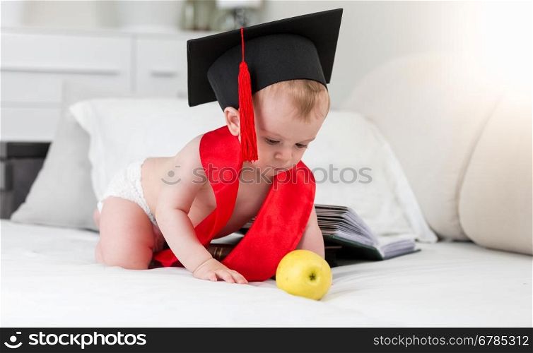 Prodigy baby in graduation cap and red ribbon reaching for apple