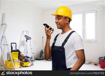 profession, building and repair concept - happy smiling indian repairman or builder in helmet using voice command recorder on smartphone over empty room with construction equipment background. happy indian builder recording voice on smartphone