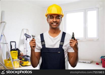profession, building and repair concept - happy smiling indian repairman or builder in helmet with wrench and pliers over empty room with construction equipment background. indian builder in helmet with wrench and pliers