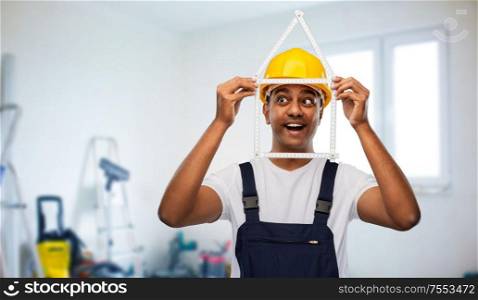 profession, building and repair concept - happy smiling indian repairman or builder in helmet with folding ruler in shape of home over empty room and construction equipment background. happy indian builder with ruler in shape of home
