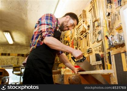 profession, carpentry, woodwork and people concept - happy smiling carpenter working with saw and wood plank at workshop