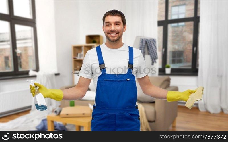 profession, cleaning service and people concept - happy smiling male worker or cleaner in overall and gloves with rag and detergent over home living room background. male cleaner with rag and detergent cleaning home