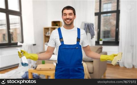 profession, cleaning service and people concept - happy smiling male worker or cleaner in overall and gloves with rag and detergent over home living room background. male cleaner with rag and detergent cleaning home
