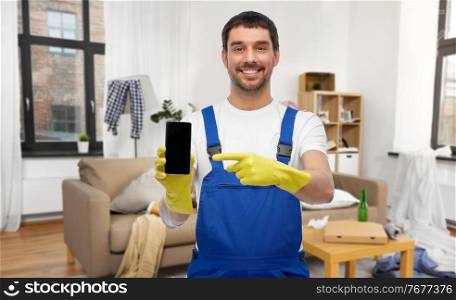 profession, cleaning service and people concept - happy smiling male worker or cleaner in overall and gloves showing smartphone over home room background. male worker or cleaner showing smartphone at home