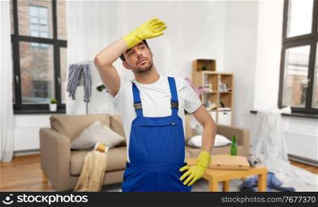 profession, cleaning service and people concept - tired male worker or cleaner in overall and gloves over home room background. tired male worker or cleaner in gloves at home