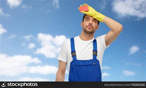 profession, cleaning service and people concept - tired male worker or cleaner in overall and gloves with sponge over blue sky and clouds on background. tired male cleaner with sponge