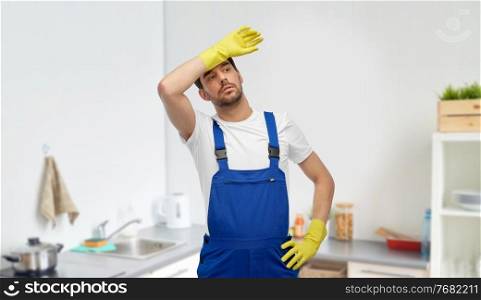 profession, cleaning service and plumbing concept - happy smiling male worker, plumber or cleaner r in overall and gloves over home room background. tired male worker or cleaner in gloves at kitchen