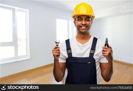 profession, construction and building concept - happy smiling indian repairman or builder in helmet with wrench and pliers over empty room background. indian builder in helmet with wrench and pliers
