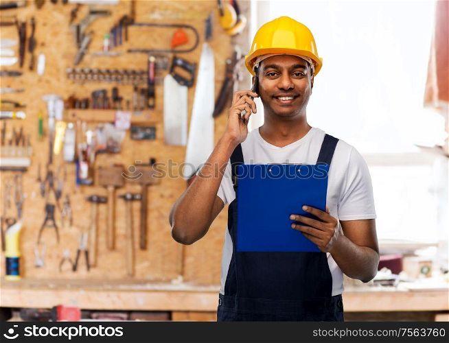 profession, construction and building concept- happy smiling indian worker or builder in helmet with clipboard calling on smartphone over work tools on background. indian builder with clipboard calling on cellphone