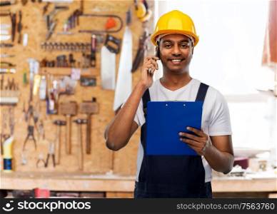 profession, construction and building concept- happy smiling indian worker or builder in helmet with clipboard calling on smartphone over work tools on background. indian builder with clipboard calling on cellphone