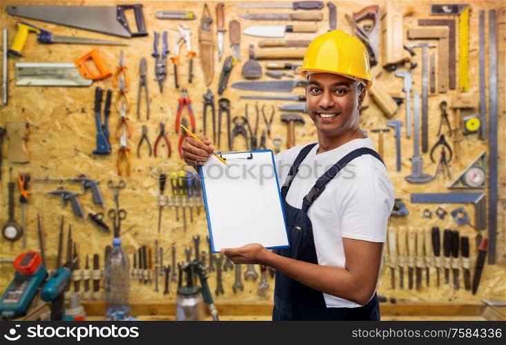 profession, construction and building concept- happy smiling indian worker or builder in helmet with clipboard and pencil over work tools on background. happy builder in helmet with clipboard and pencil
