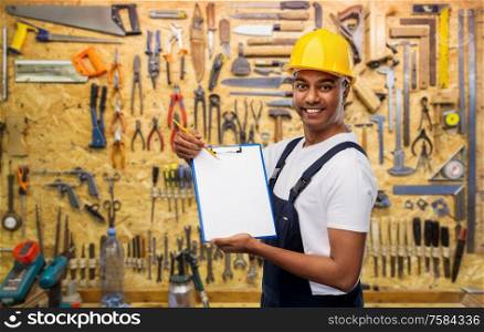 profession, construction and building concept- happy smiling indian worker or builder in helmet with clipboard and pencil over work tools on background. happy builder in helmet with clipboard and pencil