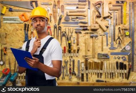 profession, construction and building concept- thinking indian worker or builder in helmet with clipboard and pencil over work tools on background. thinking builder with clipboard and pencil