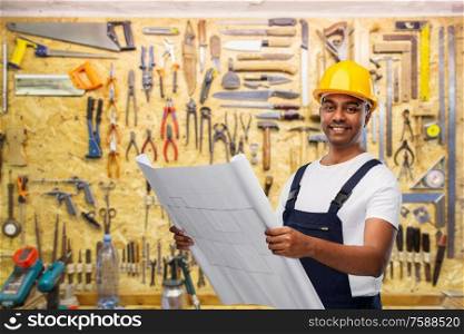 profession, construction and building - happy smiling indian builder in helmet with blueprint over working tools on wall at workshop background. happy indian builder in helmet with blueprint