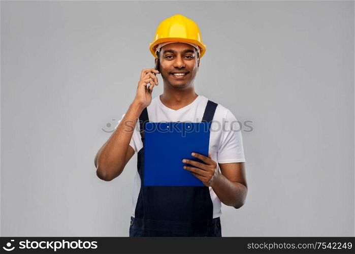 profession, construction and building - happy smiling indian worker or builder in helmet with clipboard calling on smartphone over grey background. indian builder with clipboard calling on cellphone