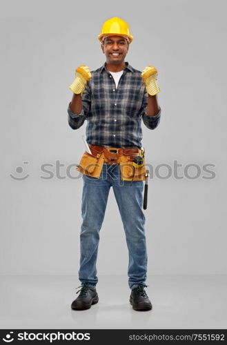 profession, construction and building - happy smiling indian worker or builder in helmet and gloves with tool belt over grey background. indian worker or builder in helmet and gloves